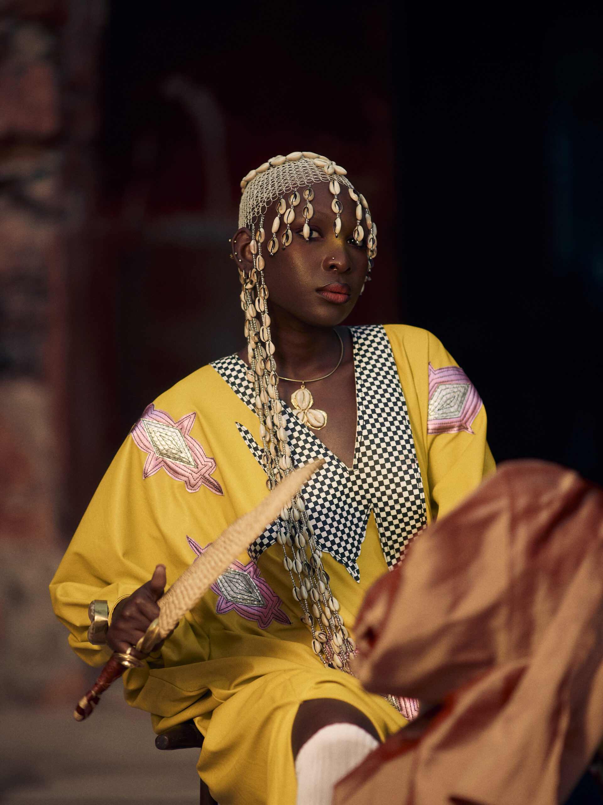 Woman holding a woven fan, wearing a yellow mboubou with checkerboard details and a cowrie-shell headpiece in the Gorée courtyard.