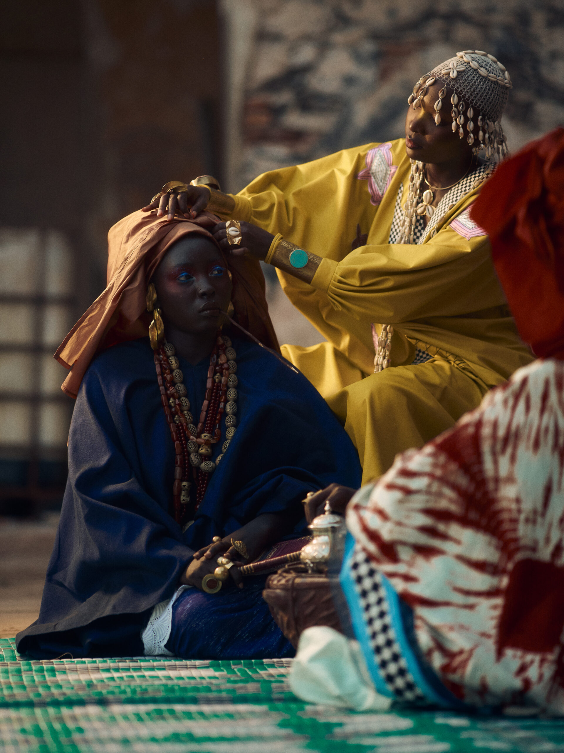 Woman in a yellow mboubou and cowrie-shell headpiece adjusts the rust-gold moussor of a seated woman dressed in indigo.