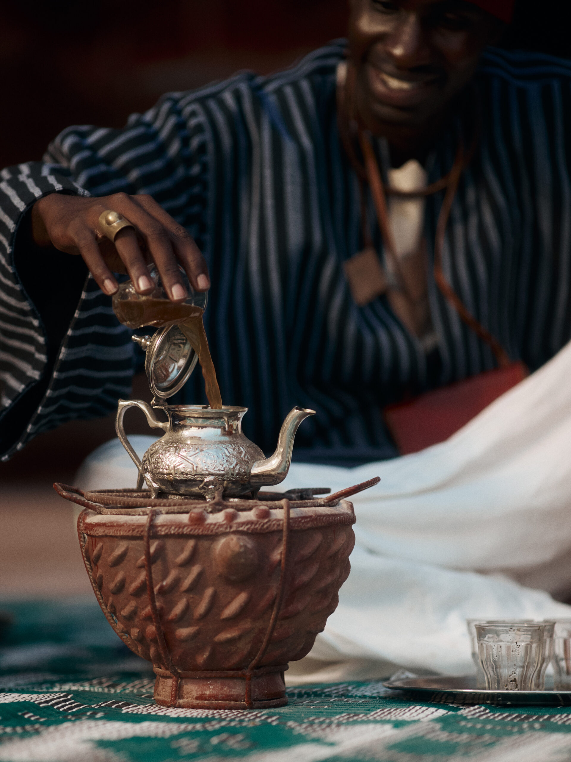 Close view of the tea host pouring attaya from a glass into an engraved silver teapot resting on a clay brazier.