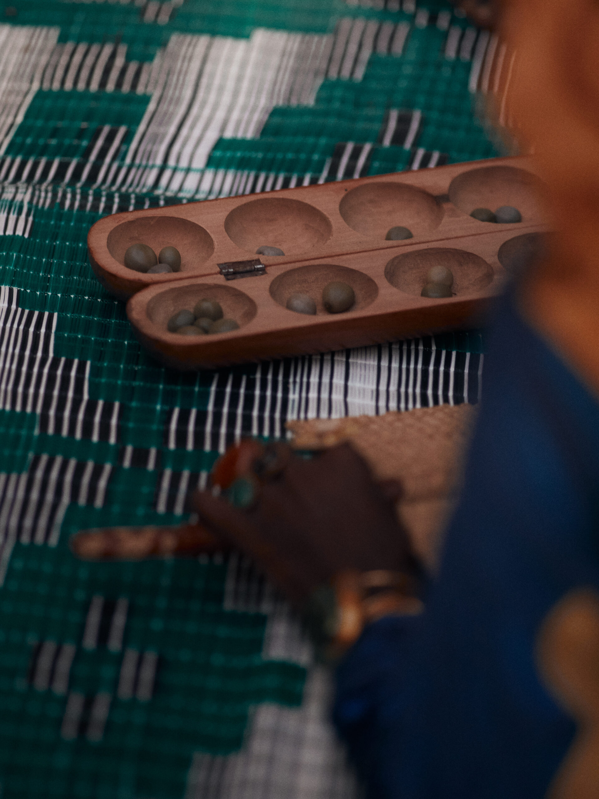 Wooden awalé board with playing pieces on a green-and-white woven basang, with a hand and woven fan in the foreground.