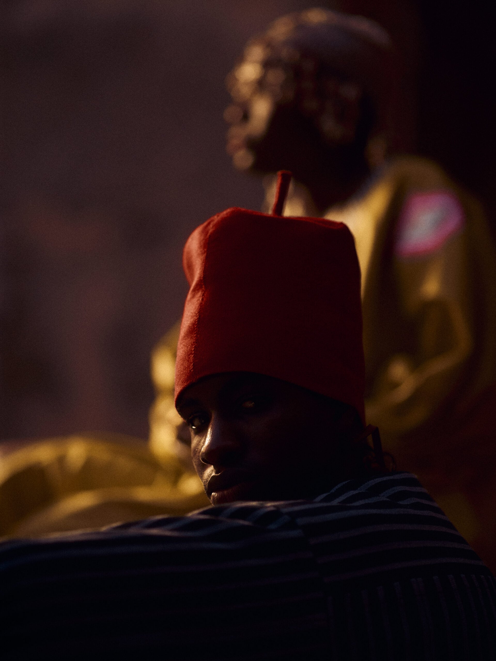 Portrait of a man in a tall red cap and blue-striped robe, his face in shadow, with a woman in yellow softly focused behind him.