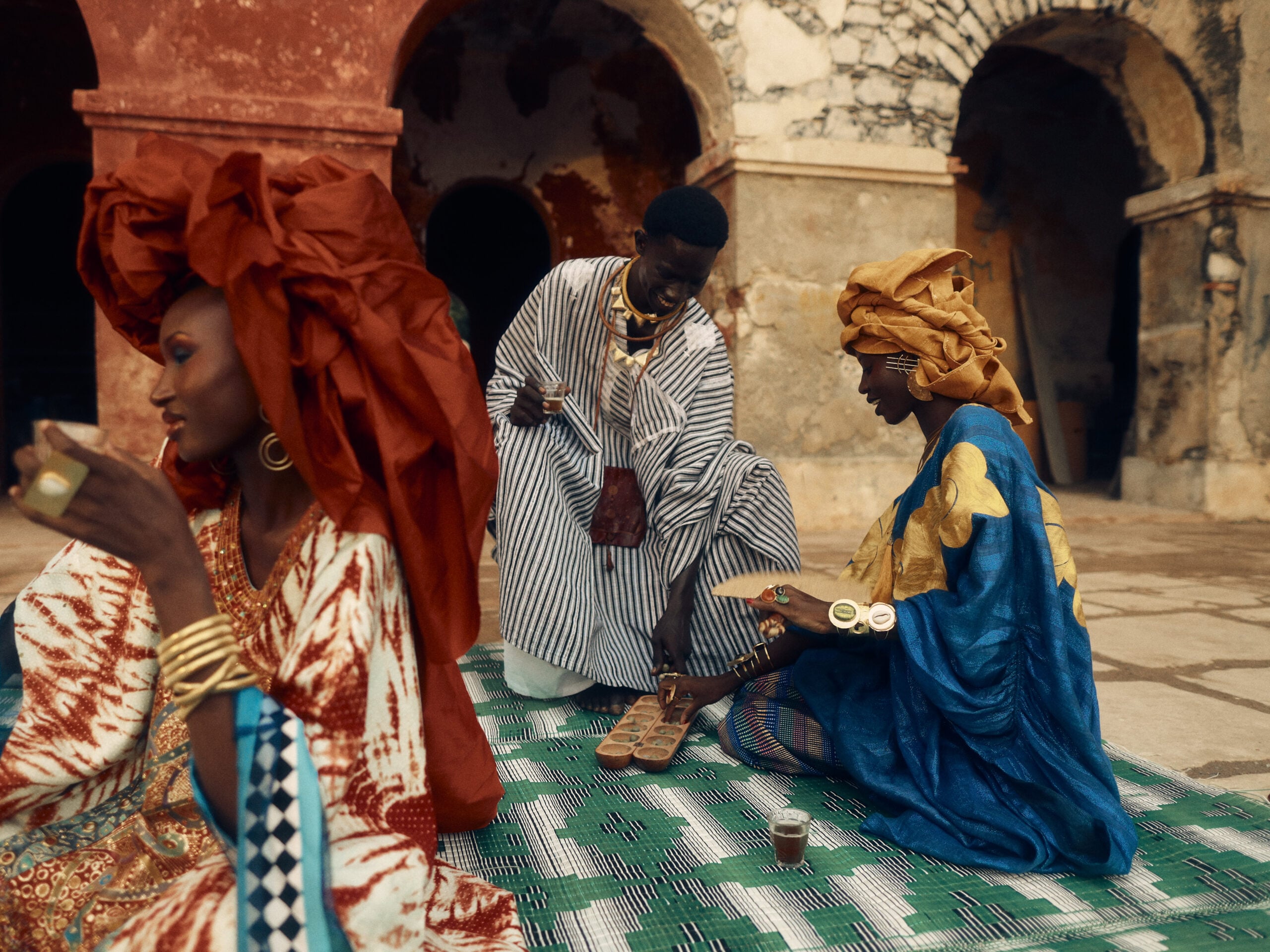 Three people gather over attaya and an awalé board on a woven mat beneath the terracotta arches of the Palais du Gouverneur on Gorée.