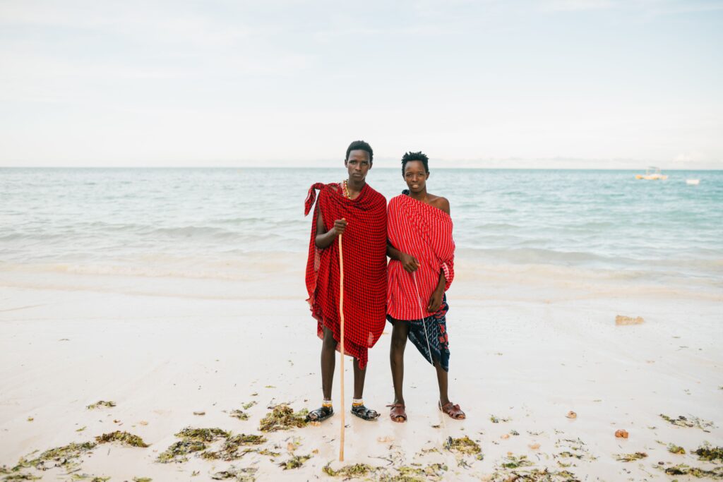 Two men wearing red patterned cloth standing together on a beach.