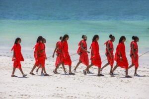 Maasai men in red shuka cloth walking along a white-sand beach beside turquoise Indian Ocean water.