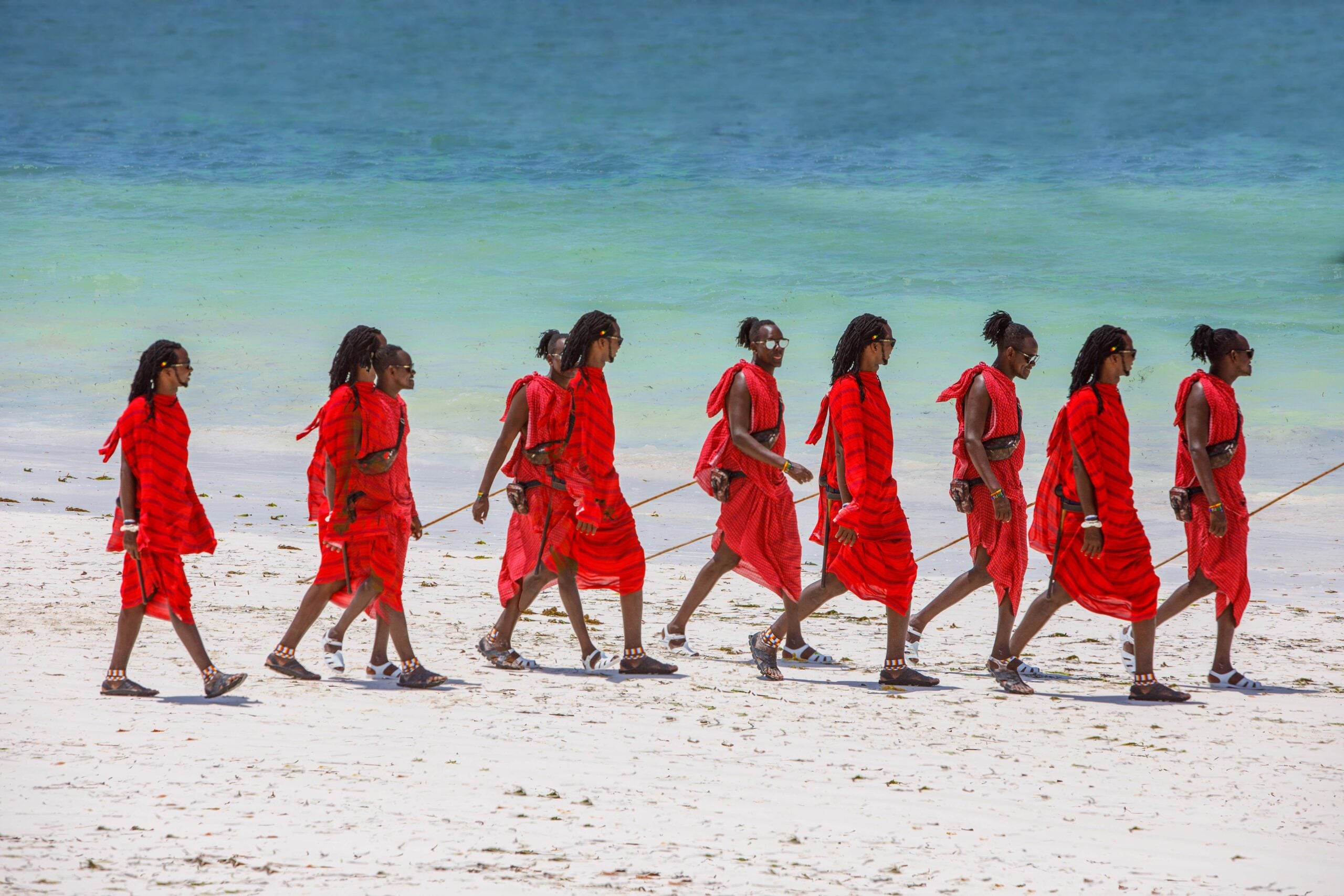 Maasai men in red shuka cloth walking along a white-sand beach beside turquoise Indian Ocean water.